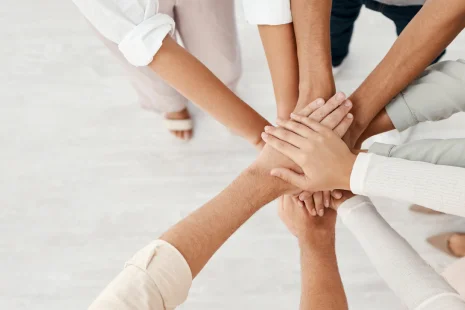 Widok z góry na różnorodne dłonie ułożone jedna na drugiej w geście jedności na jasnym tle, symbolizujący pracę zespołową, współpracę i ducha wspólnoty.